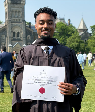 A young adult male arm amputee holds his university degree while posing outside.