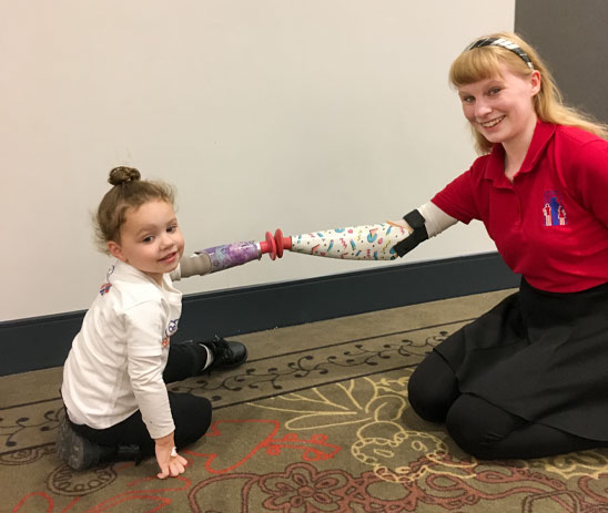 A young girl and a young woman sit on the floor and touch their artificial arms together.