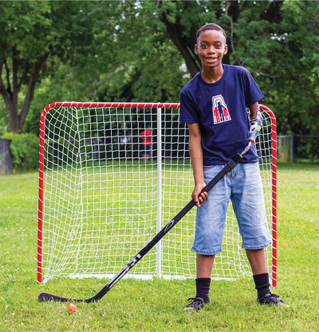 A young male arm amputee uses a hockey stick device and stands in front of a hockey net on a field.