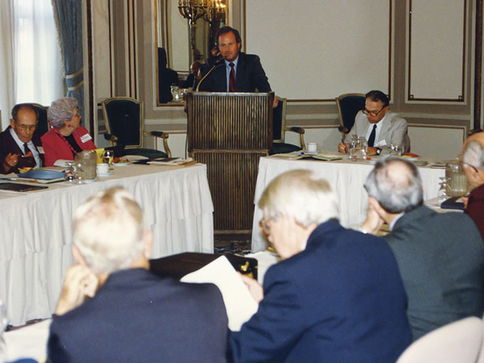 Several men and women sit at tables surrounding a podium, where a man speaks into a microphone.