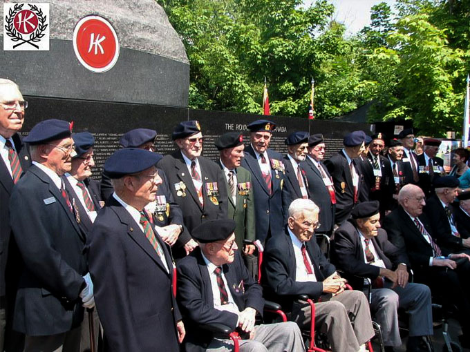 More than a dozen older amputee veterans in ceremonial dress sit and stand in front of a stone memorial wall.