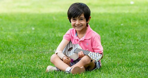 A young male arm amputee plays the ukulele while sitting in a grassy park.