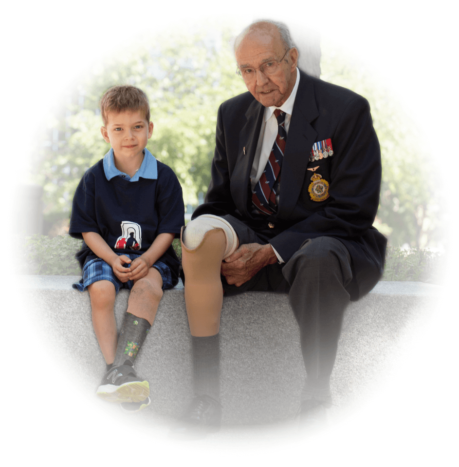 A young male leg amputee sits on a concrete wall next to an older adult male veteran with a leg amputation.