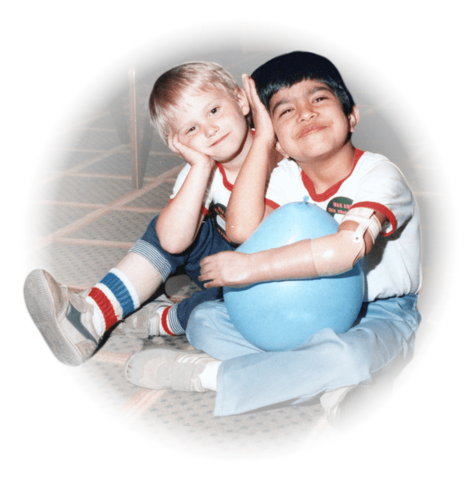  A young male arm amputee sits cross-legged on the ground holding a blue balloon next to a young male.